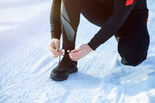 Jogging in winter. Cropped view of senior man tying laces on his sports shoes on snowy road, copy space - Photo, Image