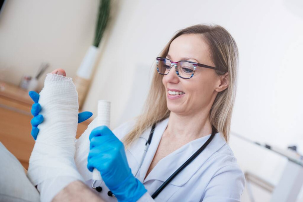 Profesjonalny uśmiech pielęgniarki bandaże nogi pacjentów z bandażem medycznym. - Zdjęcie, obraz