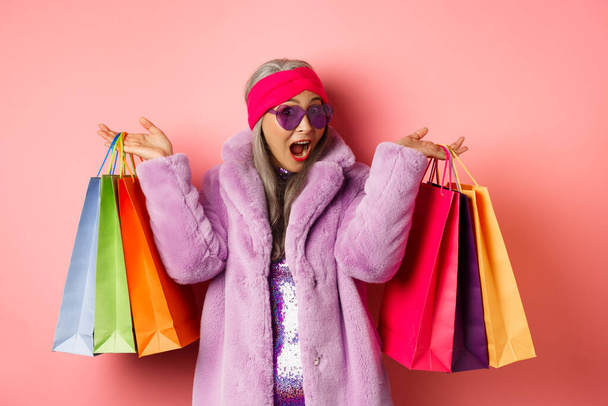 Stílusos ázsiai idős nő megy vásárolni, visel trendi ruhák és napszemüveg, holding store táskák ajándékokkal, kényeztesse magát koncepció, rózsaszín háttér - Fotó, kép