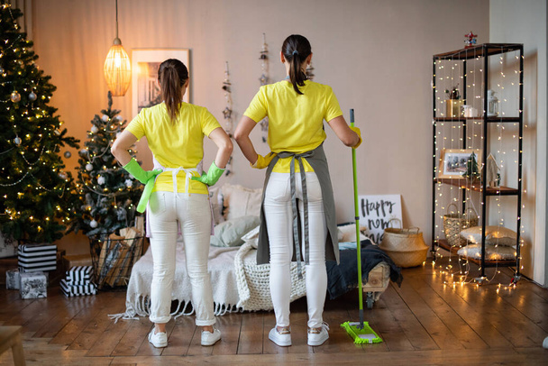 冬休み前の一般清掃クリスマスと新年。プロのハウスキーピングサービス。黄色の制服を着た二人の掃除人と手袋は腰に彼女の手で立つ - 写真・画像