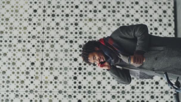 Κάθετη λήψη του νεαρού κομψό Αφρο-Αμερικανός άνδρας κρατώντας φλιτζάνι καφέ μιας χρήσης και μιλώντας στο κινητό τηλέφωνο, ενώ στέκεται σε εξωτερικούς χώρους στην πόλη την ημέρα του χειμώνα - Πλάνα, βίντεο