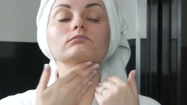 Young beautiful woman applies cream to her face in the bathroom, with a white towel on her head and in a white bathrobe. Facial skin care, prevention of early aging and wrinkles - Footage, Video