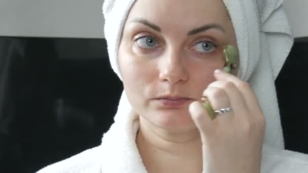 Piękna kobieta z ręcznikiem na głowie robi masaż jadeitowym wałkiem na twarz naturalnego zielonego kamienia w łazience. Chińskie narzędzia piękności. Masaż limfatyczny skóry przed starzeniem się zmarszczek - Materiał filmowy, wideo