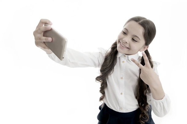 Young vlogger stream online. Child school uniform smart kid happy face. Girl cute long curly hair holds smartphone taking selfie white background. Child girl school uniform clothes holds smartphone - 写真・画像