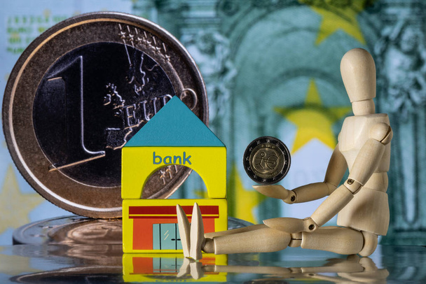 条件付き銀行の建物の近くに座っている象徴的な人の木製の図は、拡大1ユーロ硬貨と100ユーロ紙幣の断片を背景に、彼の手に2ユーロ硬貨を持っています。 - 写真・画像