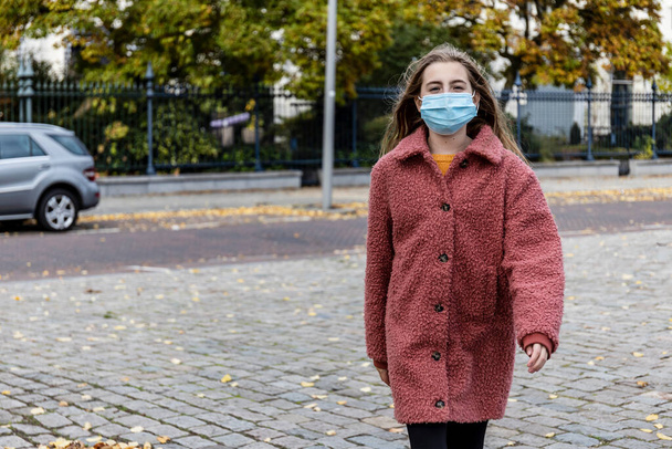 Chica rubia en abrigo de invierno rosa caminando con confianza en una calle de la ciudad con una máscara para la pandemia del virus Corona. - Foto, imagen