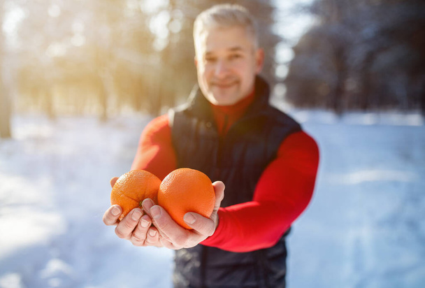 Sportivo senior che offre frutta arancione come pasto sano dopo l'allenamento nel parco invernale innevato, messa a fuoco selettiva - Foto, immagini