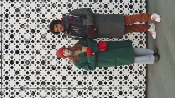 Scatto verticale della giovane donna caucasica e dell'uomo afro-americano che tengono in mano le tazze da caffè e guardano la macchina fotografica mentre stanno insieme di fronte al moderno edificio urbano nella giornata invernale - Filmati, video