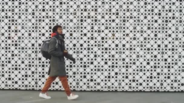 Full length tracking shot of young Afro-American man in stylish outterwear holding to go coffee and walking along modern building in city on winter day - Πλάνα, βίντεο