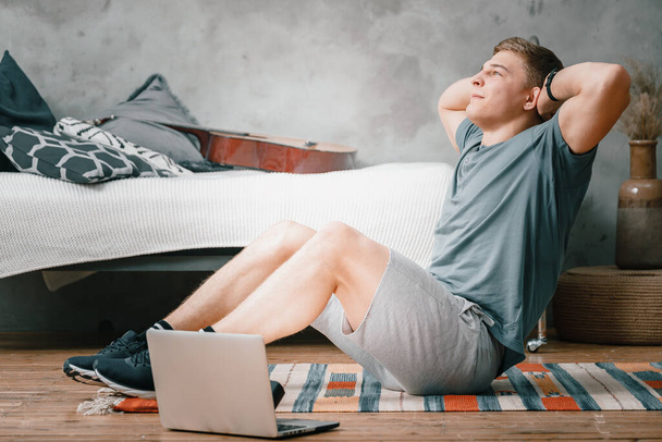 Young man goes in for sports at home, training online. The athlete makes the press, smiles, there is an open laptop, bed, vase, carpet nearby. - Photo, image
