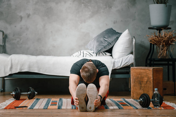 Ein junger Mann treibt zu Hause Sport, trainiert online am Telefon. Der Sportler streckt sich im Schlafzimmer, im Hintergrund ein Bett, eine Vase, ein Teppich. - Foto, Bild