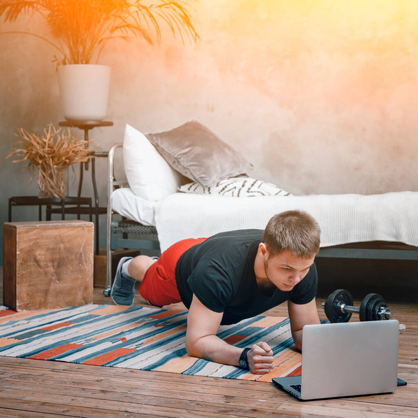 Young man goes in for sports at home, training online. The athlete makes a plank, watches a movie and studies from a laptop  in the bedroom, in the background a bed, a vase, a carpet. - Photo, Image