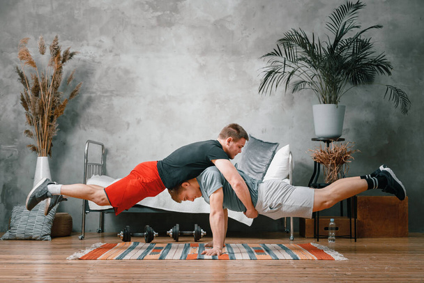 The athlete men makes a plank on top of each other in the bedroom, in the background a bed, a vase, a carpet. Gymnasts training, acrobats' home tricks  - 写真・画像