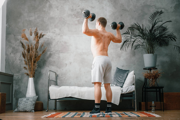 A  athlete with blond  hair shakes biceps in dumbbells in the bedroom,  online training. The young man goes in for sports at home. - Фото, зображення