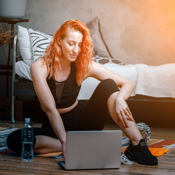 Close-up of a young woman in a sports uniform is resting on the floor at home,  watches a movie and studies from a laptop   , a social network. Student loafing around and putting off work and study - Photo, Image