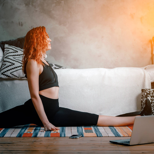 A young woman goes in for sports at home, online workout . The athlete  stretching , meditating, sitting on a twine  in the bedroom, in the background there is a bed, a vase, a carpet. - Photo, Image