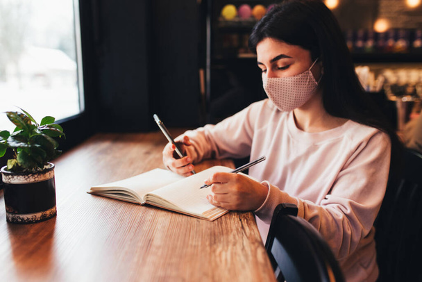 Yüz maskeli kız, telefon okuyor ve deftere yazı yazıyor. Bulanık arkaplan - Fotoğraf, Görsel