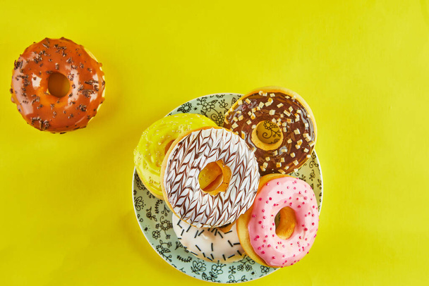 Multicolored donuts with icing and splashes on a blue plate on a yellow background. Flat lay - Photo, Image