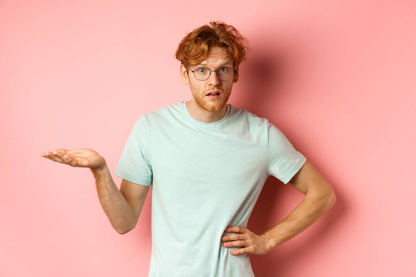 So what. Confused redhead man raising hand and shrugging, looking puzzled, dont understand something, standing over pink background - Фото, зображення