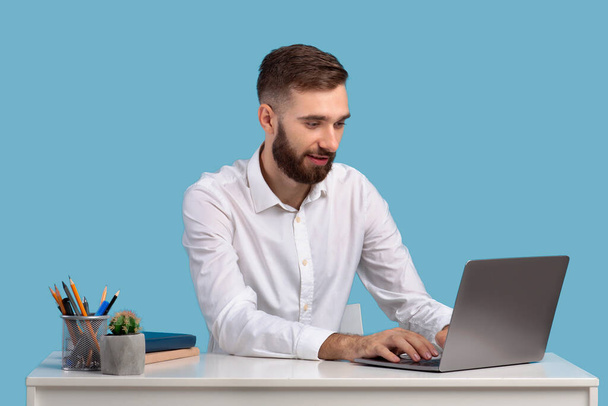 Концепция работы онлайн. Привлекательный молодой бизнесмен, используя ноутбук за своим столом на синем фоне студии - Фото, изображение