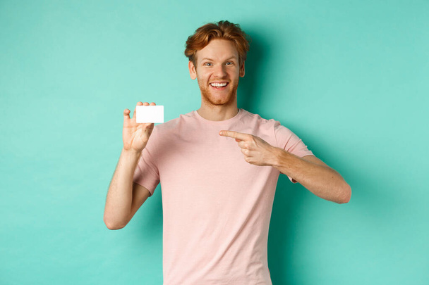 Ελκυστικός ενήλικας με γενειάδα και κόκκινα μαλλιά που δείχνουν με το δάχτυλο την πλαστική πιστωτική κάρτα, χαμογελώντας ευχαριστημένος στην κάμερα, στέκεται πάνω από τυρκουάζ φόντο - Φωτογραφία, εικόνα