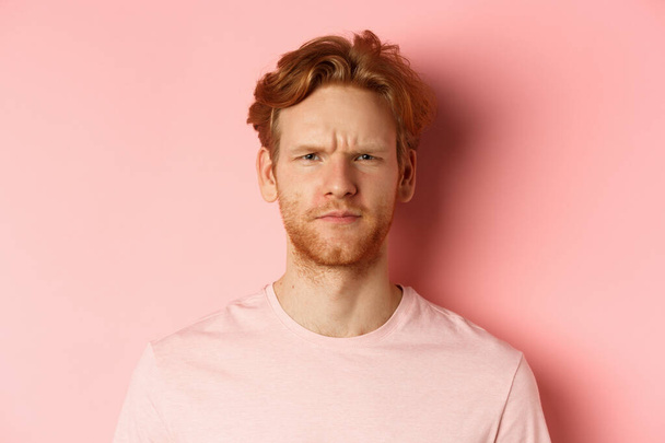 Close up of suspicious redhead guy frowning, staring at camera doubtful, express disapproval, standing over pink background - Photo, Image