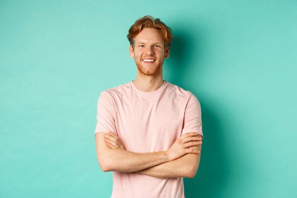 Portret van vriendelijk ogende jongeman met rood haar en baard glimlachend en starend tevreden, hand in hand gekruist op de borst, staande over turquoise achtergrond - Foto, afbeelding