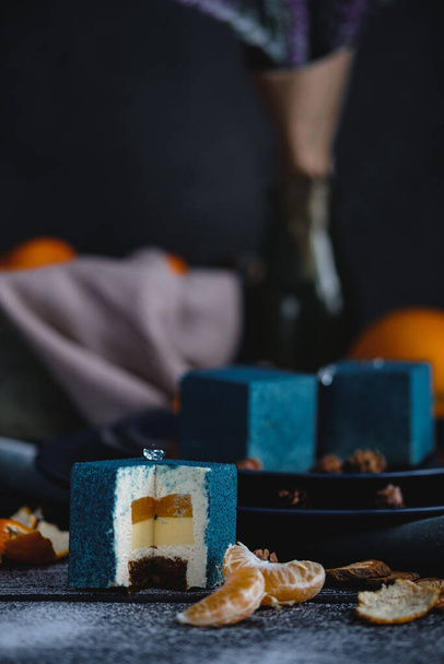 Blue cake filled with orange and tangerine in the shape of a cube. Nearby on a black table are ingredients oranges, tangerines, nuts. Rustic style - Фото, изображение