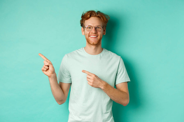Kısa kızıl saçlı ve gözlüklü neşeli genç adam, parmaklarını fotokopi alanına gösteriyor, gülümseyen beyaz dişler, reklam gösteriyor, nane arkaplanı. - Fotoğraf, Görsel