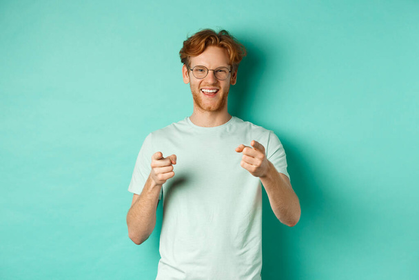 Przystojny młodzieniec z rudymi włosami, noszący okulary i koszulkę, wytykający palcami kamerę i uśmiechający się, wybierający ciebie, gratulujący lub chwalący, stojący nad miętowym tłem - Zdjęcie, obraz