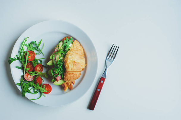 healthy tasty breakfast - croissant with avocado, arugula, ham and cheese served with cherry tomato salad on white plate - Photo, Image
