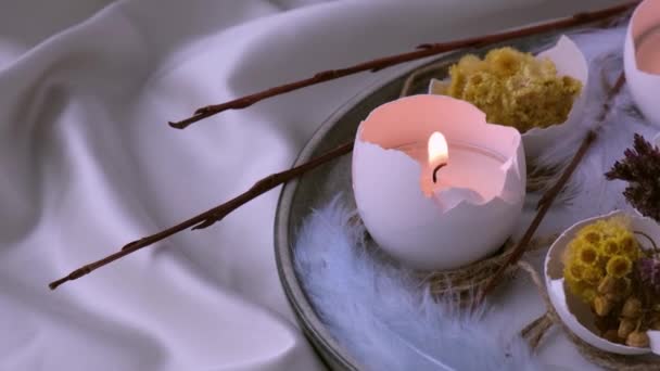 Модные пасхальные украшения. Разбитые органические яйца с сухими травяными цветами на тарелке. Свечи в белой скорлупе яиц. Птичье перо. Пылающие свечи пламенный дым - Кадры, видео