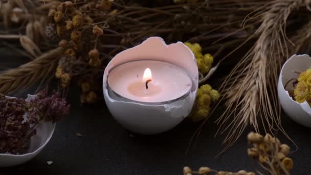 Burning candle Trendy Easter decorations. Cracked organic eggs shell with dry grass and dry flowers candle light. Dark background. - Footage, Video