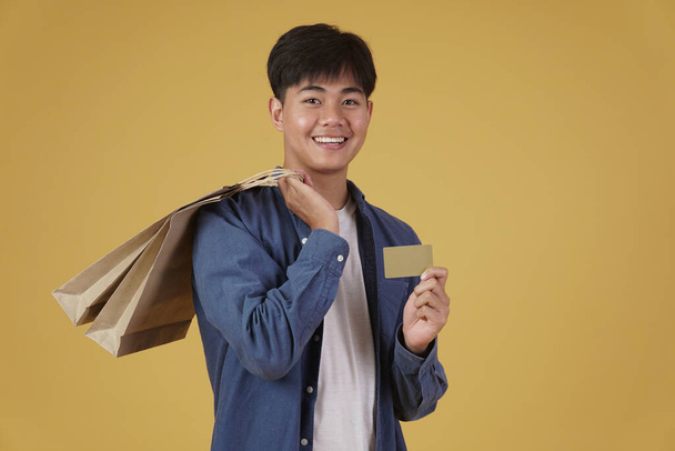 πορτρέτο του νεαρού ευτυχισμένου Ασιάτη άνδρα ντυμένος άνετα κρατώντας τσάντες για ψώνια και πιστωτική κάρτα που απομονώνονται σε κίτρινο φόντο στούντιο - Φωτογραφία, εικόνα