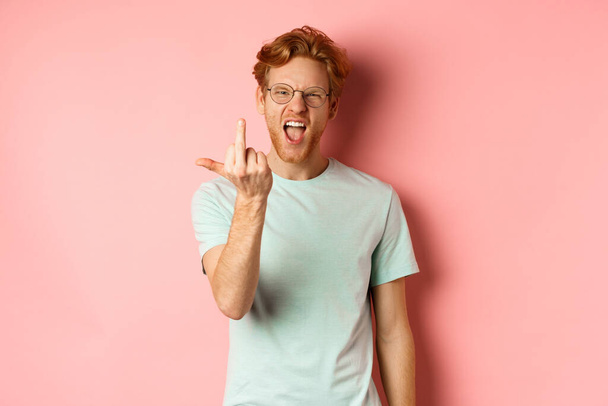 Arrogant and rude redhead man in glasses dont give a fuck, showing middle fingers at camera and frowning, standing over pink background - Фото, изображение