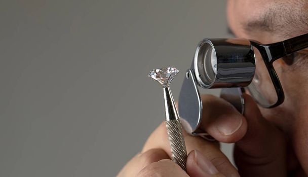 Asian man with glasses diamond dealer buyer expert evaluates polished diamond through magnifying glass close up with copy space. - Foto, afbeelding