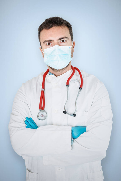Ο γιατρός άνθρωπος πορτρέτο απομονώνονται σε μπλε φόντο φορώντας μάσκα προσώπου με τα χέρια σταυρωμένα - Φωτογραφία, εικόνα