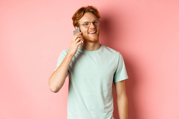 Bellissimo ragazzo hipster con i capelli rossi e la barba che parla sul cellulare, chiama qualcuno e sembra felice, in piedi su sfondo rosa - Foto, immagini