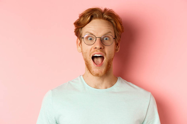 Close up of surprised redhead man raising eyebrows and scream with joy, checking out cool promo, looking amazed at camera, standing over pink background - Φωτογραφία, εικόνα