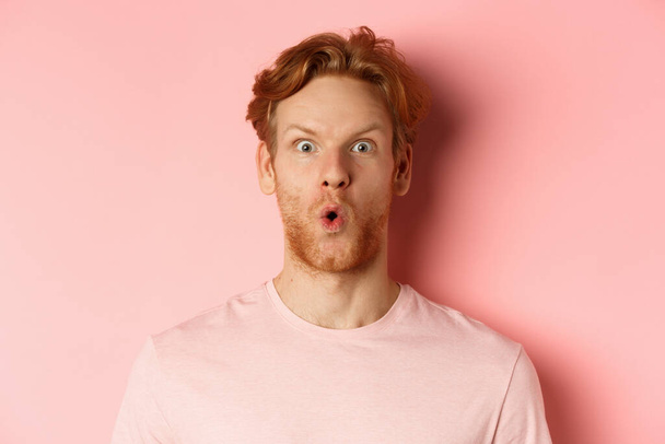Headshot van de jonge roodharige man met baard, zeggen wow en staren naar camera verbaasd, het controleren van promo deal, staande over roze achtergrond - Foto, afbeelding