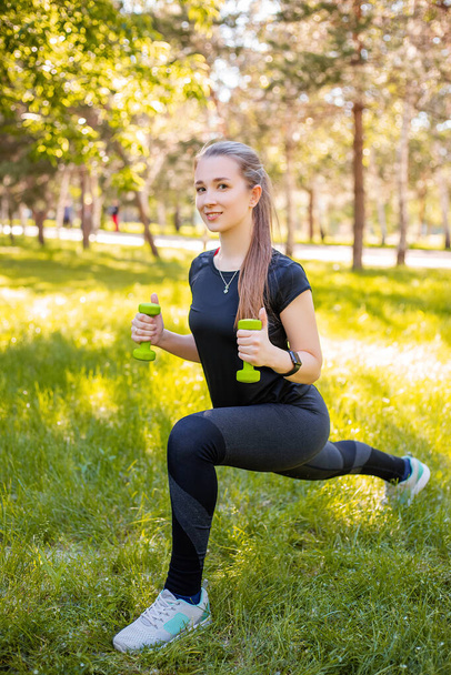 An attractive girl in a sports uniform makes a lunge exercise to stretch the muscles of the legs with dumbbells in her hands. The concept of independent fitness training in nature. - Zdjęcie, obraz