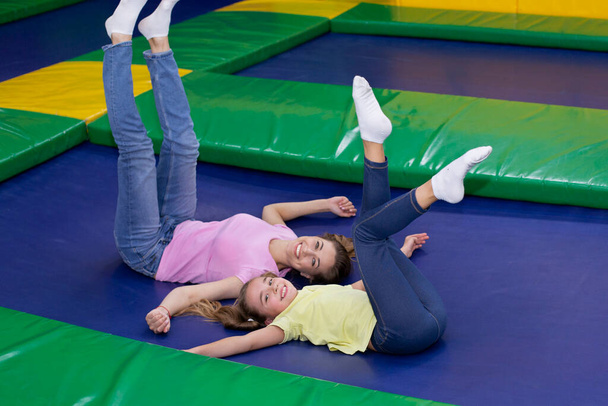 Cute teen girl and her happy mother lying on trampoline, lifting their legs up, having fun together at amusement park - Photo, Image