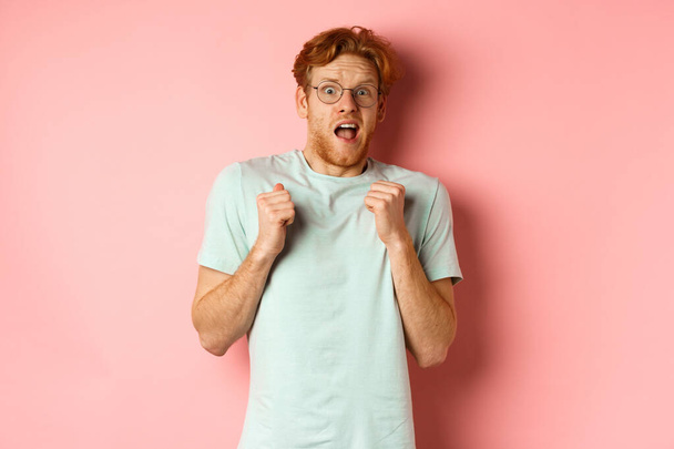 Portrait of scared redhead guy in glasses staring startled at camera, press hands to body and scream of fear, standing over pink background - Photo, Image