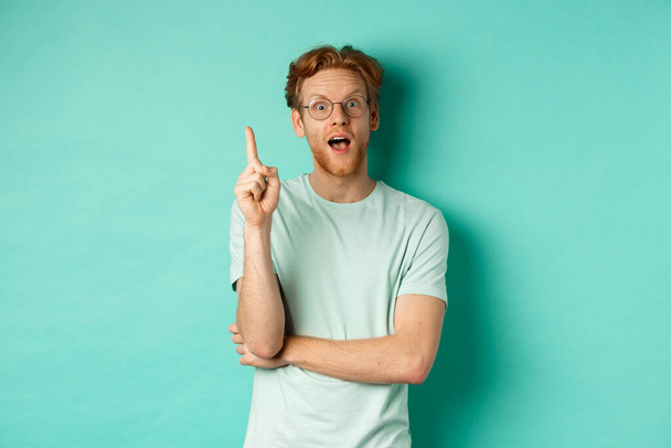 Возбужденный молодой человек с рыжими волосами в очках, поднимающий указательный палец, подающий идею, стоящий на мятном фоне - Фото, изображение