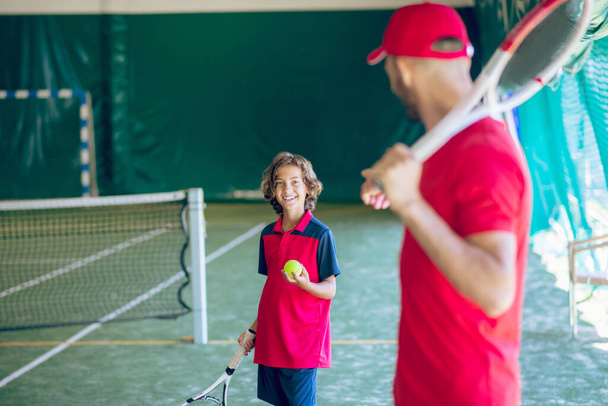 Young bearded man in a red cap holding a tennis racket and talking to a boy - Фото, зображення