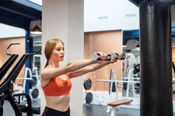 Beautiful young woman trains deltoid in gym with dumb-bell - Photo, image