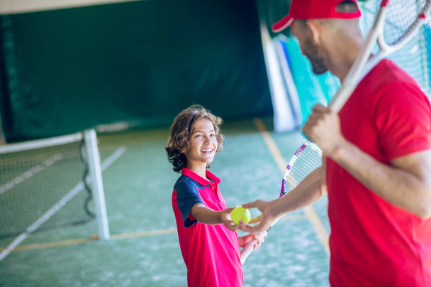 Young bearded man in a red cap shaking hands with a dark-haired boy before playing tennis - Photo, image
