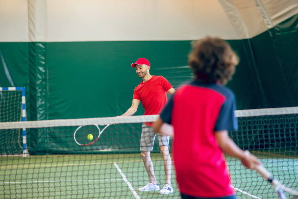 Coach in red clothes and cap playing tennis with a boy - Фото, зображення