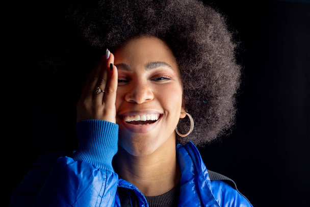 hezká smíšená afro žena, usmívající se, s městským oblečením zakrýt půlku obličeje s rukou, tmavé pozadí - Fotografie, Obrázek