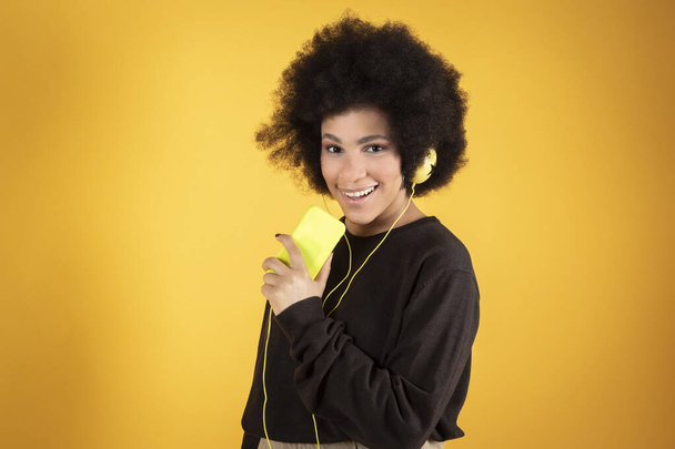 gemengde afro vrouw, met koptelefoon en smartphone, zingende gele achtergrond - Foto, afbeelding